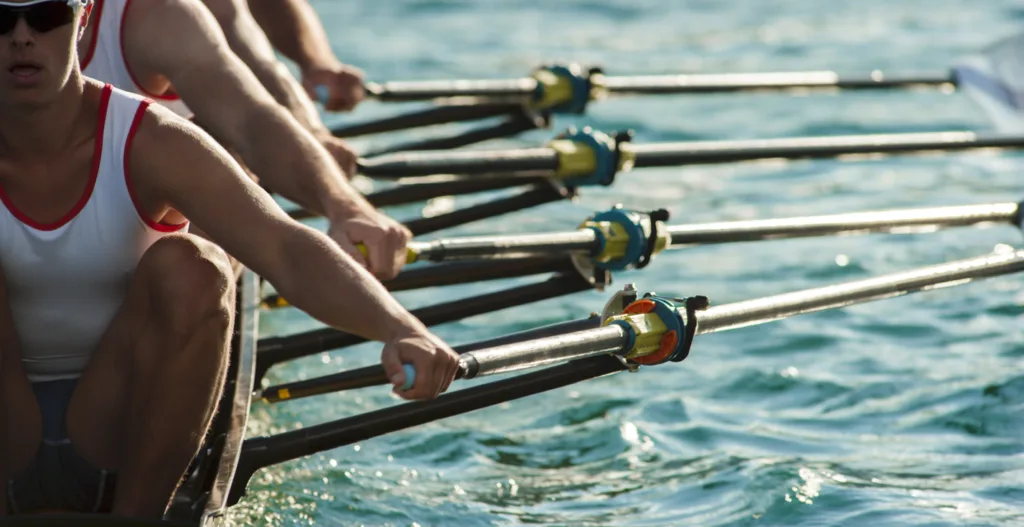 A close-up side view of a rowing team in action, with a focus on the athletes' upper bodies and oars. The team members are wearing matching tank tops, part of a coordinated crew. Their arms are extended, gripping the oars, which are positioned parallel to each other and dipped into the water, indicating synchronized movement. The sunlight reflects off the water, creating a shimmering effect, and highlights the muscles and focus of the rowers, emphasizing the physical effort and teamwork involved in the sport.